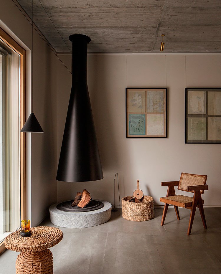 Chimenea circular de hierro, base elevada de hormigón, suelo cerámico, cesto y mesilla de fibra natural, butaca de madera