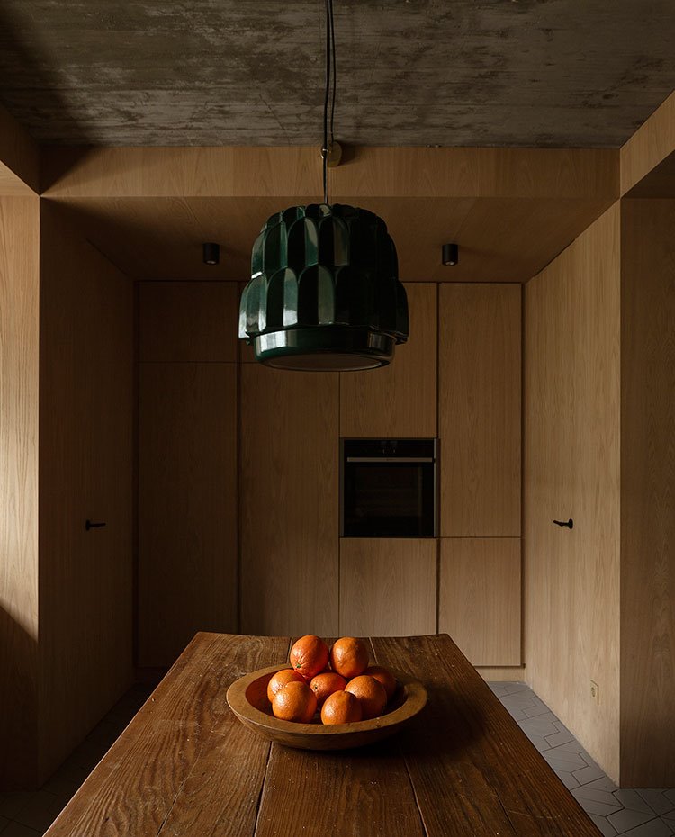 Cocina con mesa de madera, centro de mesa con naranjas, luminaria suspendida en verde oscuro, frentes de armario en madera y techo de hormigón