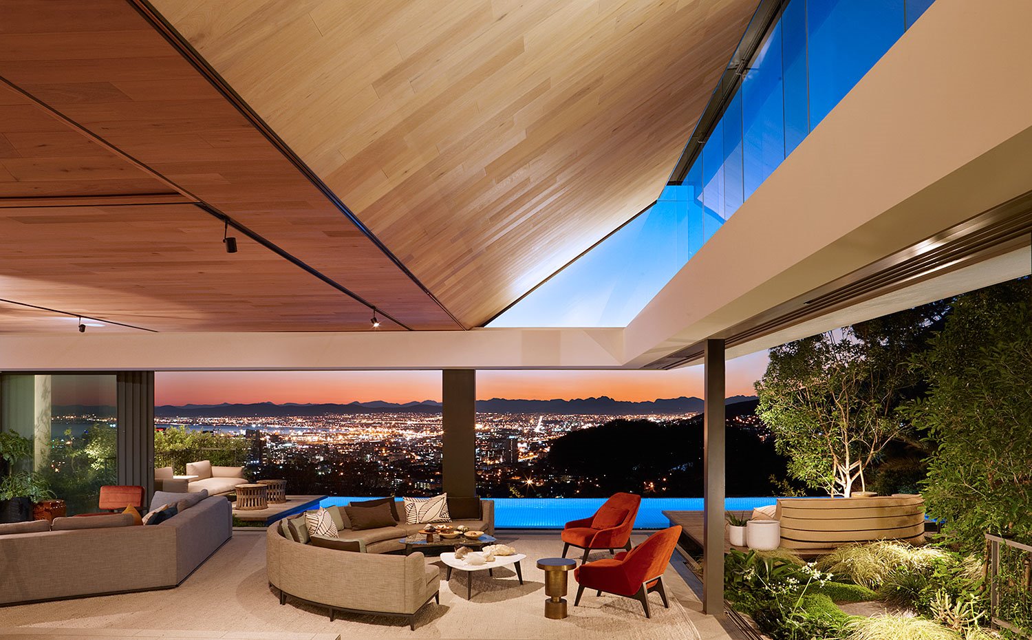 Salón con sofá semicircular, mesa de centro blanca y negra y butacas naranjas frente a piscina asomando al skyline nocturno de la ciudad