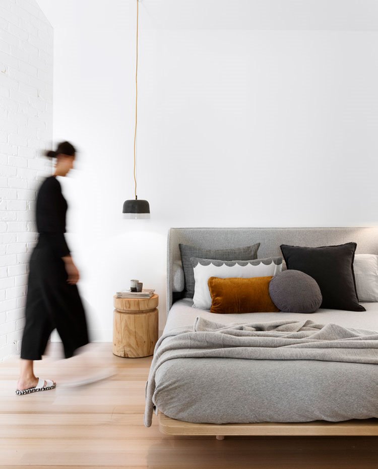 Dormitorio con paredes blancas y suelo de madera, cama con estructura de madera y cabecero gris, lámpara suspendida de techo sobre mesilla de noche en madera