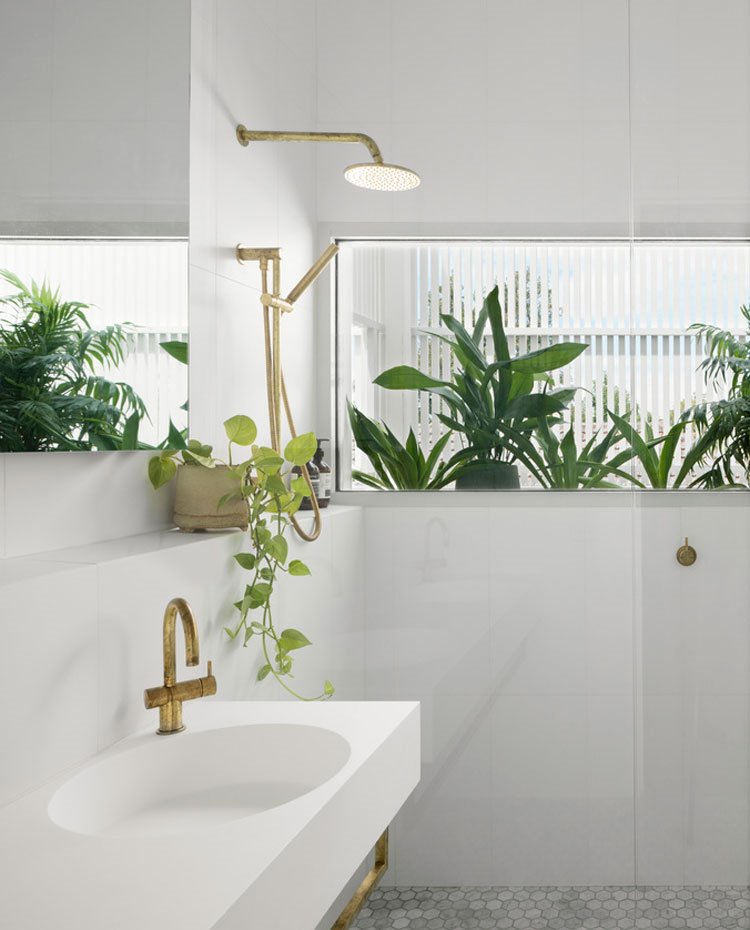 Lavamanos blanco con grifería cromada dorada, revestimiento en blanco, plantas como elementos divisorios hacia ducha abierta