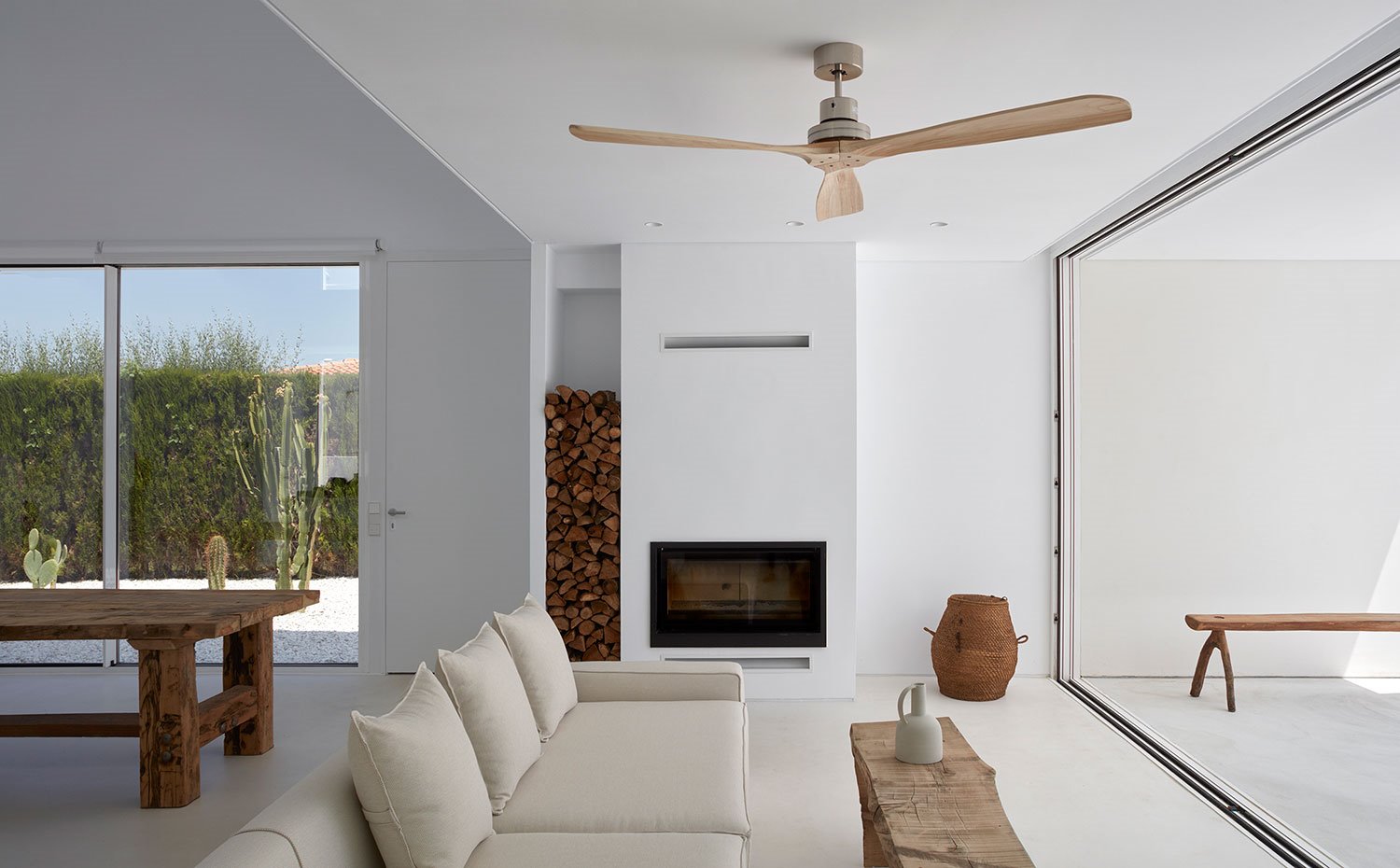 Mesa comedor, cerramiento de cristal, chimenea de obra con hueco para troncos, sofá beige, ventilador de techo de madera