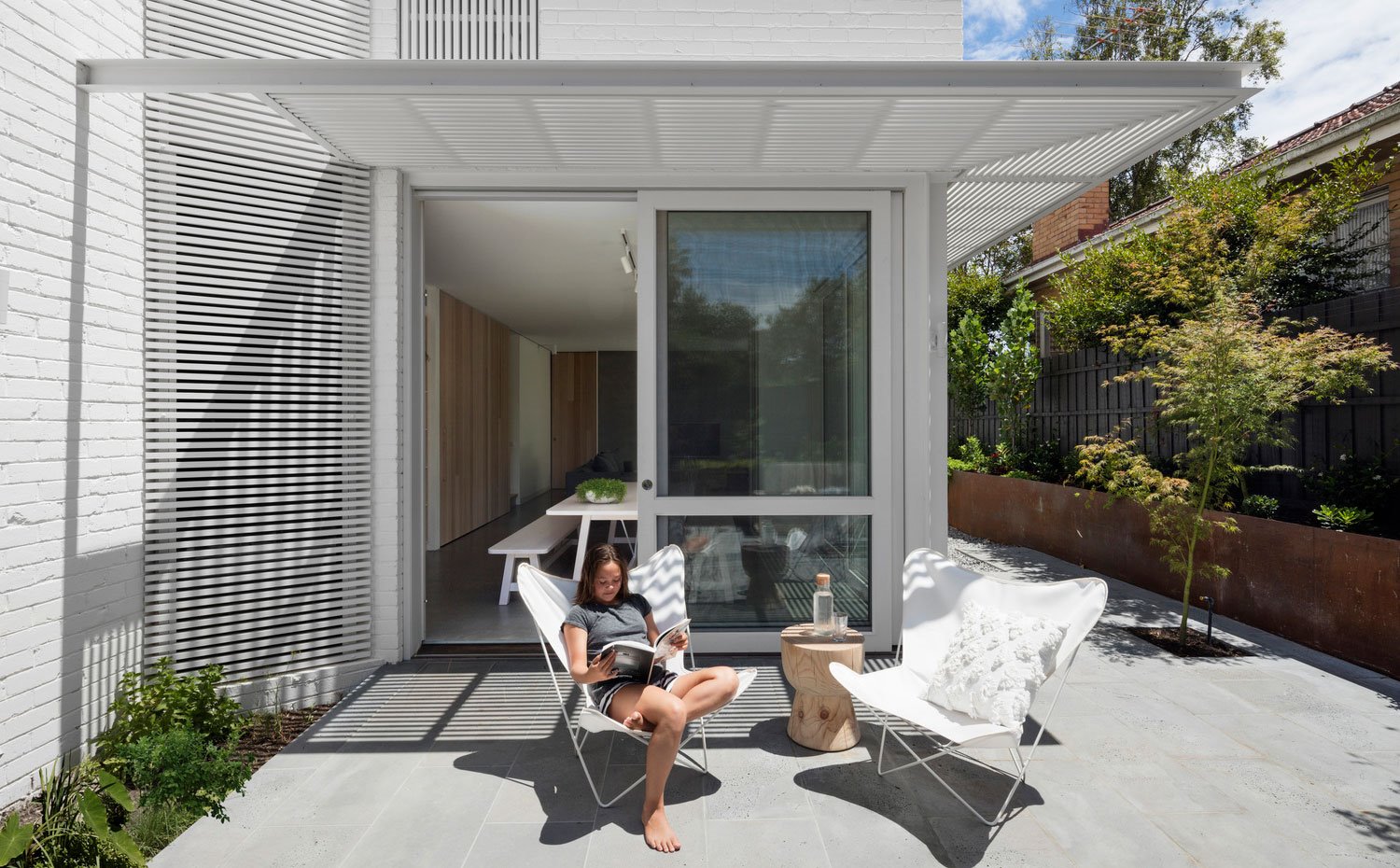 Patio exterior con butacas en blanco y mesita auxiliar de madera, cerramiento de aluminio con mesa y bancada de madera blanca
