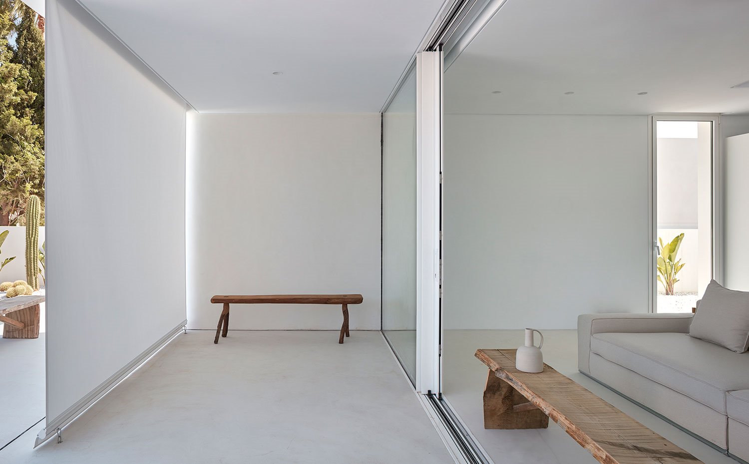 Salón orientado a exterior con bancos de madera, sofá beige, suelo continuo, lona separadora de ambientes, exterior con vegetación