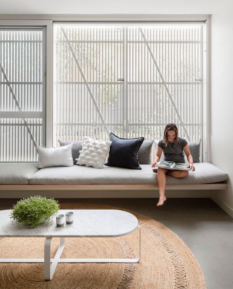 Zona de estar con mesa de sobre de granito y estructura en blanco, alfombra de fibra natural, sofá hecho a medida con estructura de madera y cojines en gris, blanco y negro, cerramiento con celosías blancas