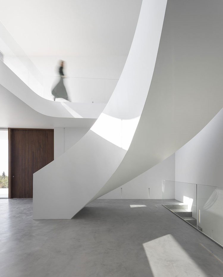 Espacio a doble nivel, escalera curva en blanco sobre pavimento en hormigón, barandilla de cristal