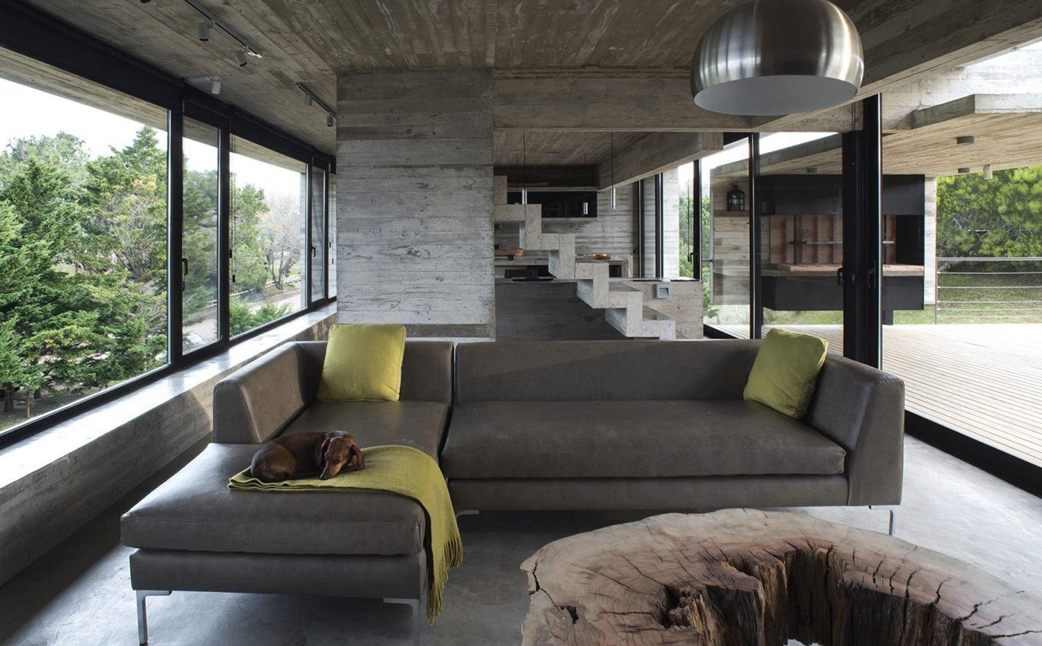Zona de estar con sofá gris, cojines y manta pistacho, estructura de hormigón con cocina abierta al fondo