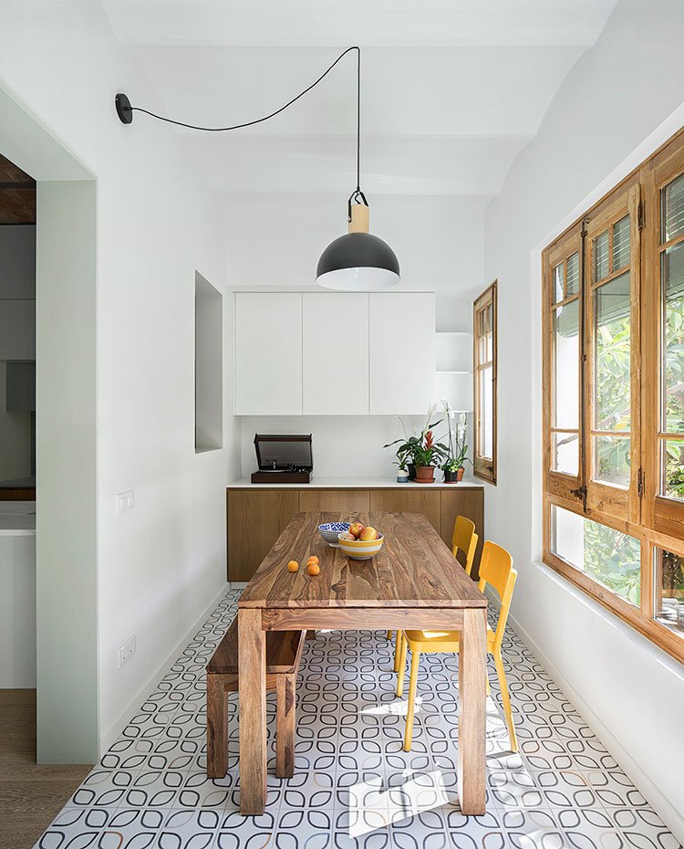 Comedor con mesa y banco de madera y sillas en amarillo, luminaria de suspensión con patalla en negro y detalles dorados, mueble almacenaje en pared en blanco y madera
