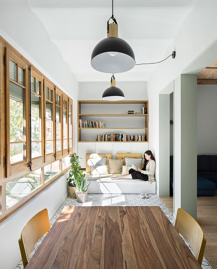 Vista frontal rincón de lectura con mesa de comedor de madera y sillas amarillas, luminarias suspendidas, mueble almacenaje en pared y asiento acolchado tapizado con motivos amarillos