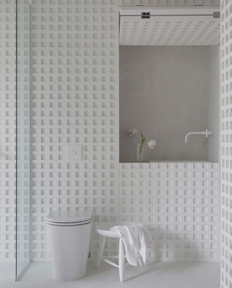 Detalle lavamanos de baño de habitación e inodoro, con grifería en blanco, lavamanos en hueco abierto junto a jarrón con flores blancas y estructura perforada con motivos geométricos