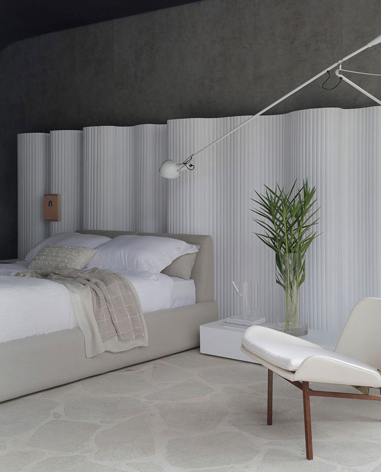 Dormitorio con trasera con una gran estructura blanca de formas curvas, butaca blanca con acabado de amdera, ropa de cama de tonos crudos, suelo de piedra