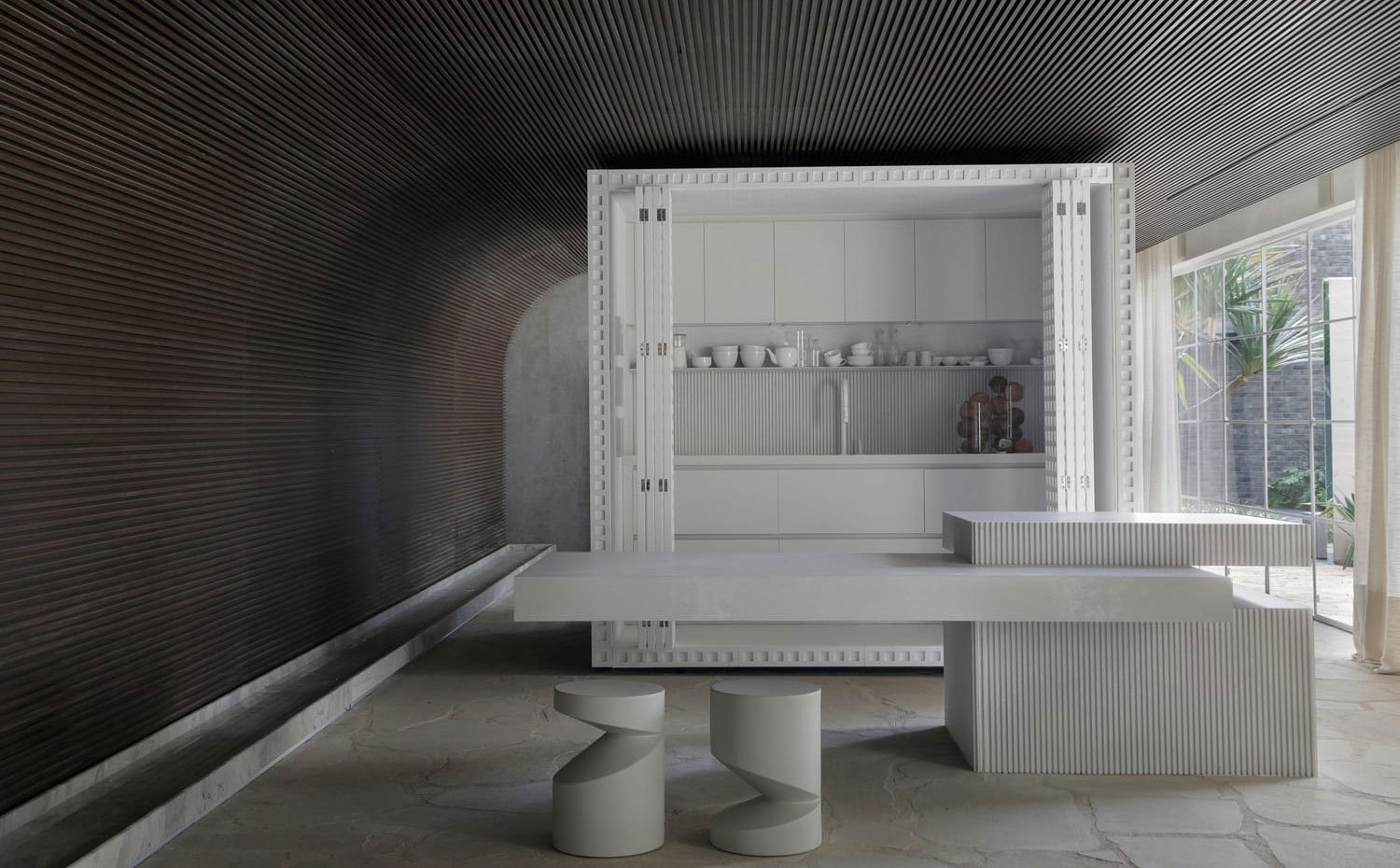 Módulo blanco con cocina integrada y puertas plegables, isla de trabajo con base blanca alistonada y encimera volada para office, dos taburetes blancos