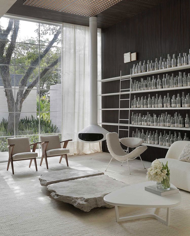 Salón con estantería de botellas de cristal, chimenea suspendida, butacas, sofá y mesa de centro en blanco, cerramiento acristalado con cortina blanca de techo a suelo