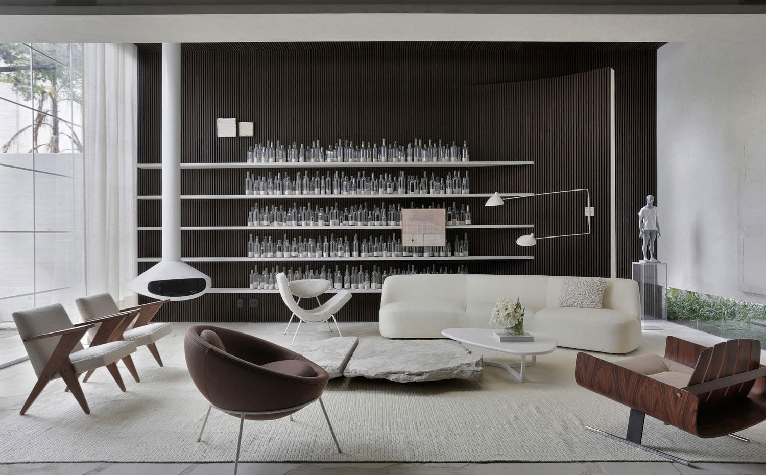 Salón con sofás y butacas en blanco y madera, alfombra en blanco, chimenea suspendida blanca, baldas con botellas sobre pared alistonada, cerramiento de cristal con cortinas blancas
