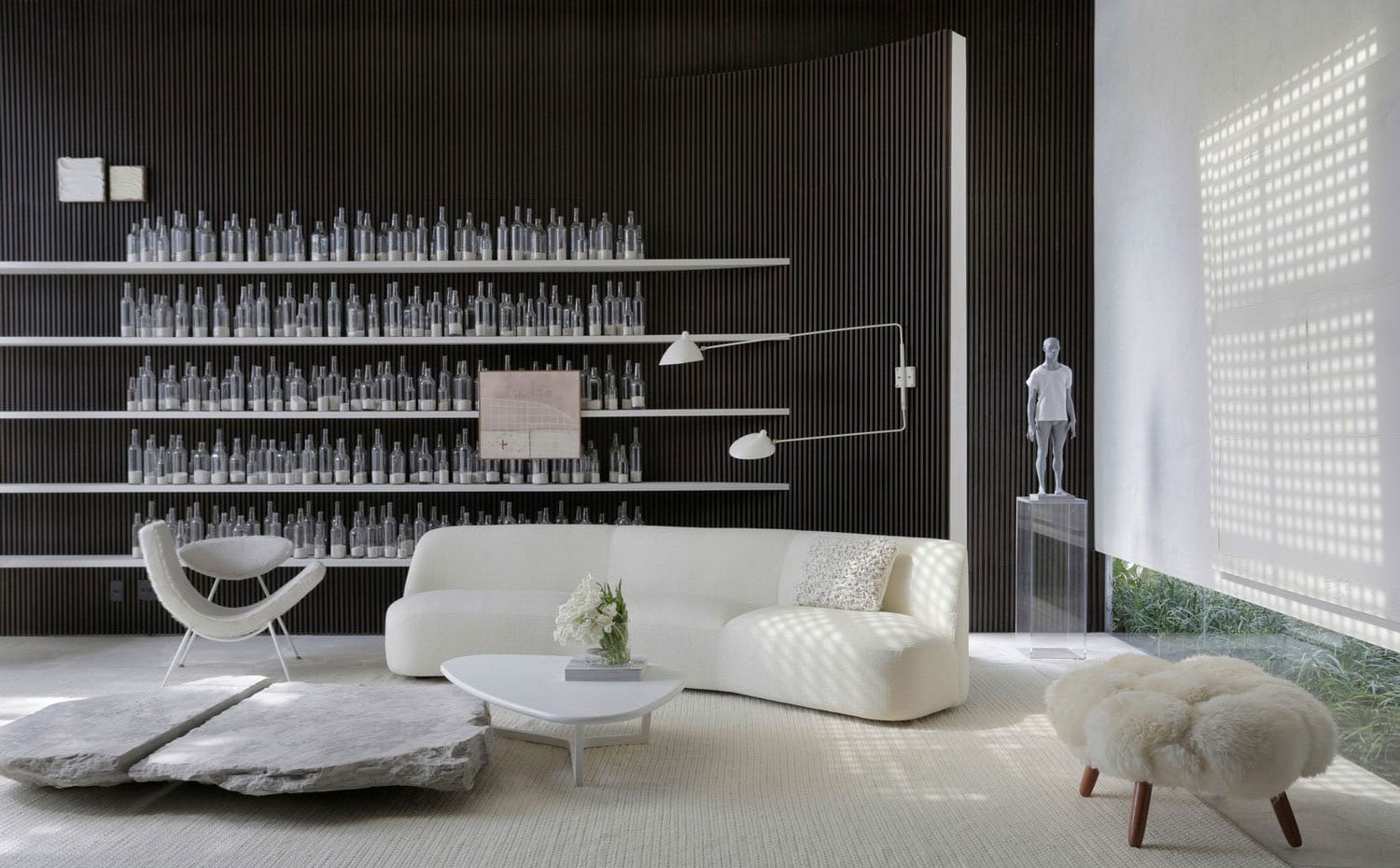 Zona de estar con mobiliario en blanco, estantería con botellas de cristal, escultura en gris sobre pedestal de metacrilato, pared alistonada