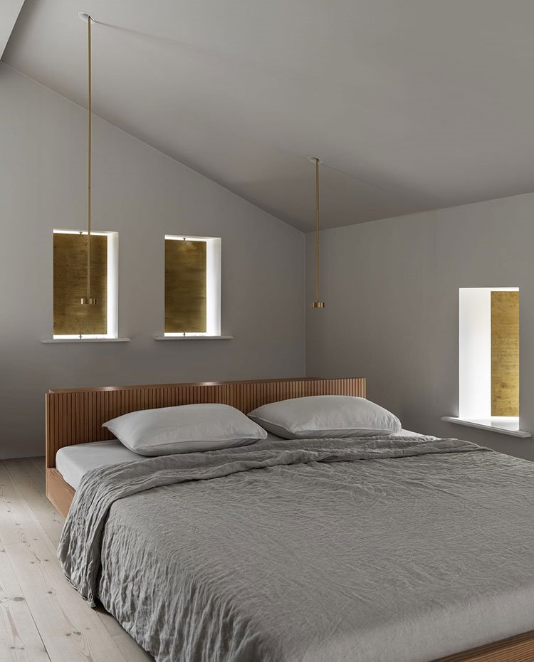 Dormitorio con cabecero de cama en madera, luminarias suspendidas doradas, sábana gris, suelo de madera, cerramientos ventanas dorados