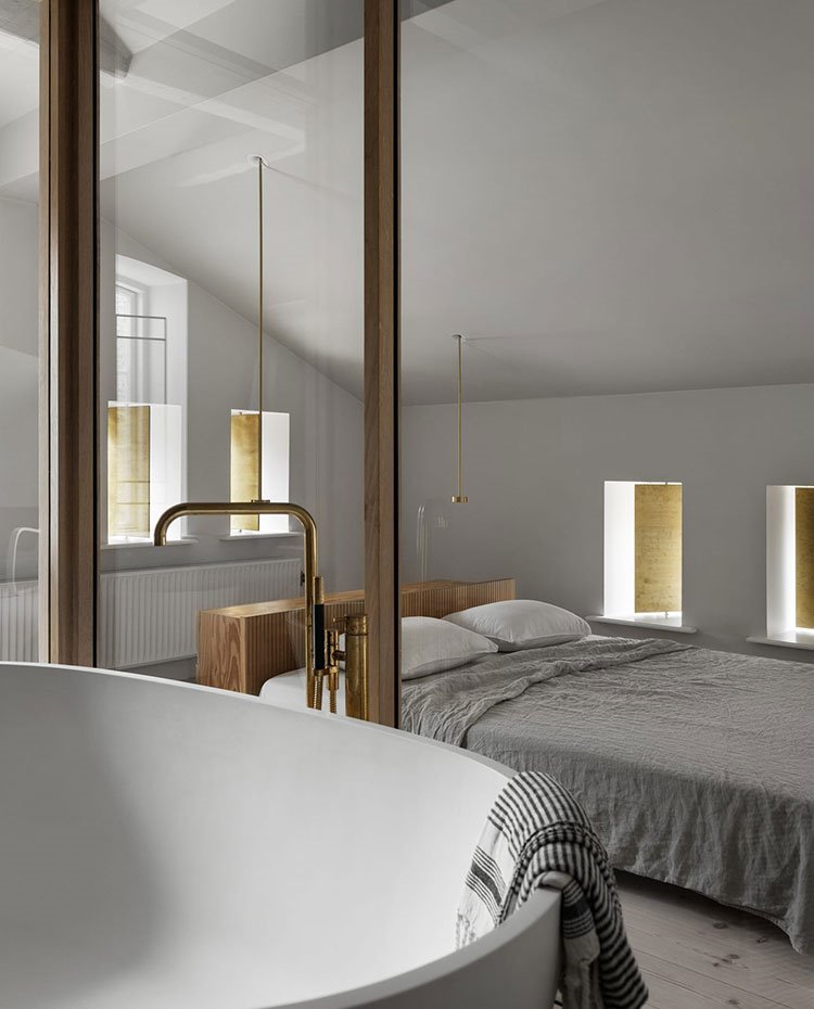 Estructura de cristal y perfilería de aluminio de cuarto de baño, bañera exenta, grifería dorada, cama dormitorio al fondo