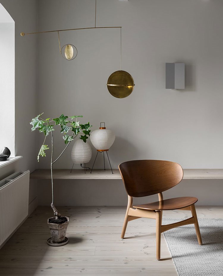 Zona de estar con silla de madera, alfombra en beige, balda suspendida junto a pared con dos lámparas de sobremesa, planta en el suelo y detalles escultórico dorado