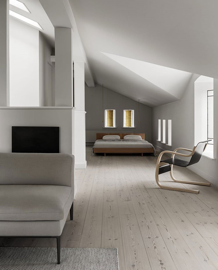 Zona de estar junto a dormitorio con sofá y alfombra en color beige, suelo de madera, butaca negra con estrutura de madera junto a ventana y cama de dormitorio