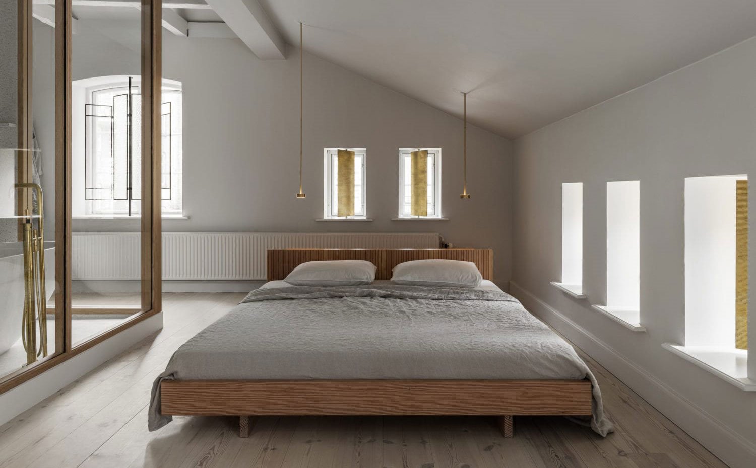 Zona dormitorio, cama con estructura de madera, luminarias suspendidas junto a cabecero, cerramiento de cristal con cuarto de baño