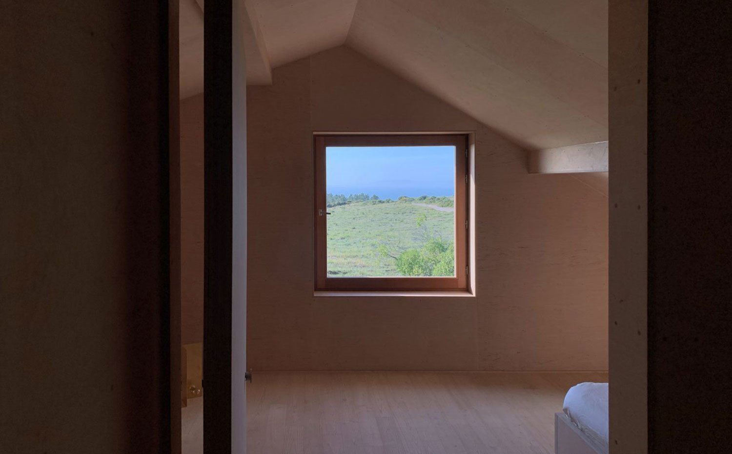 Detalle dormitorio con techo inclinado a dos aguas y ventana cuadrada a paisaje exterior, todo revestido de madera