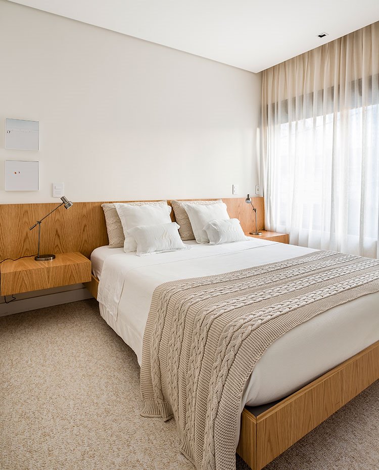 Dormitorio con estructura de cabecera, mesitas y cama en madera, manta beige sobre cama, cortinas blancas