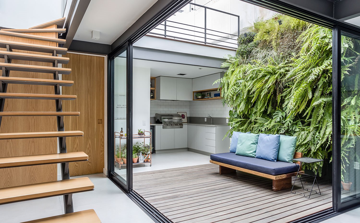 Patio interior con jardín vertical, banco con tapicería en azul, barbacoa semicubierta, escaleras de madera hacia planta superior