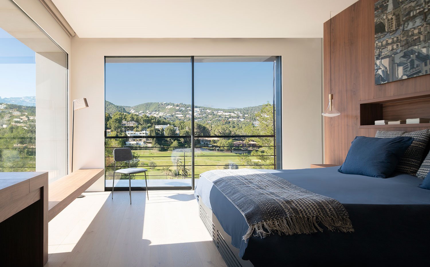 Dormitorio con pared de cabecero revestida de madera, ropa de cama en azul petróleo, mesita con bancada en perímetro de pared, cerramiento de cristal