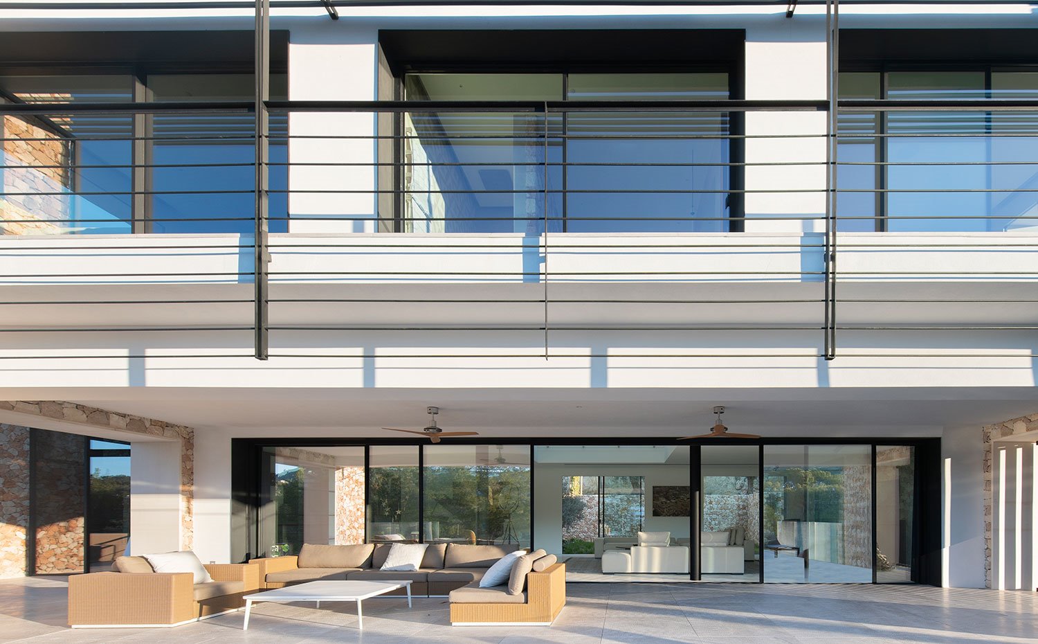 Porche exterior con sofás de fibra trenzada y gran mesa de centro, cerramientos de cristal a salón y balcones de nivel superior 