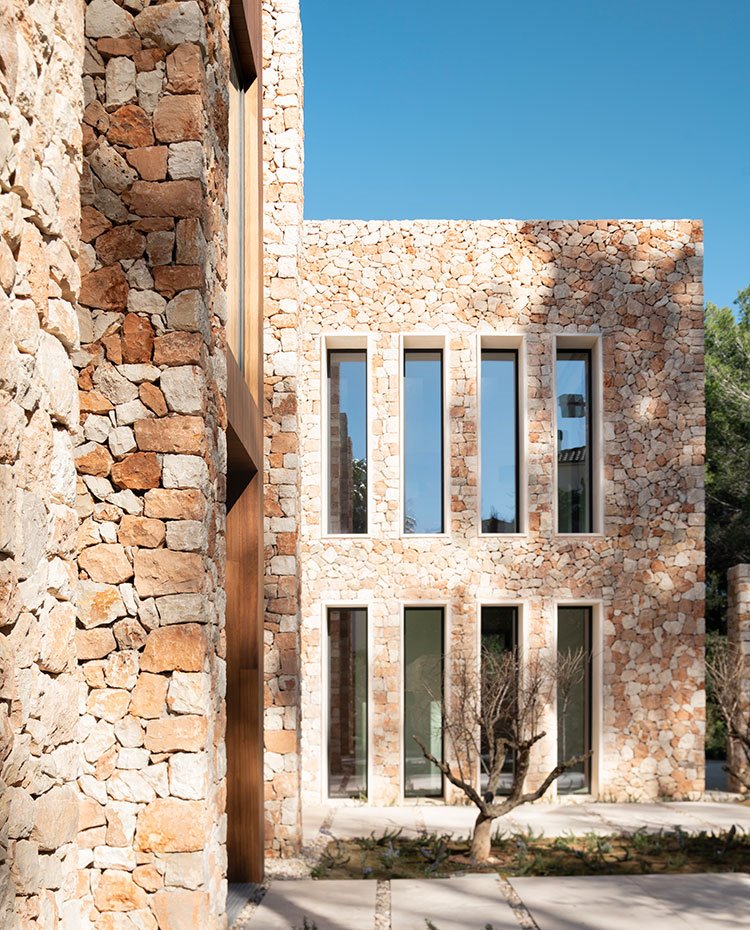Secuencia de ventanas verticales junto a la puerta de entrada con muros de piedra y árboles frutales