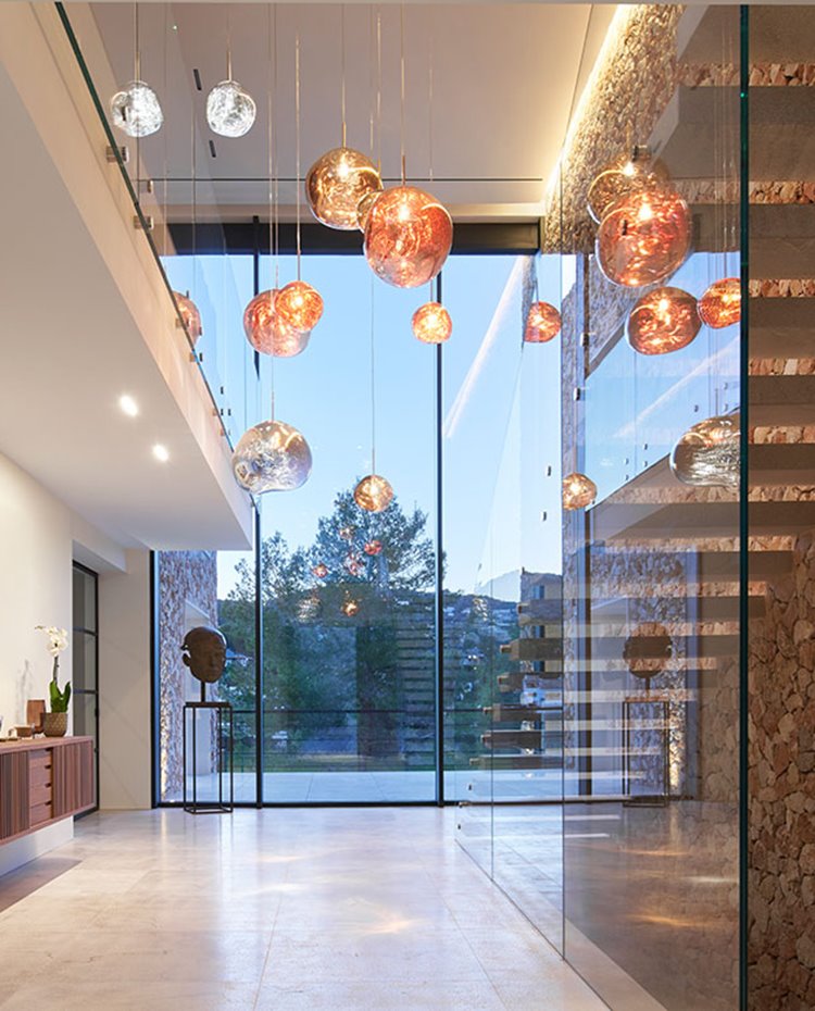 Zona de paso con hueco de escalera revestida de cristal transparente, mueble suspendido y luminarias supendidas a gran altura