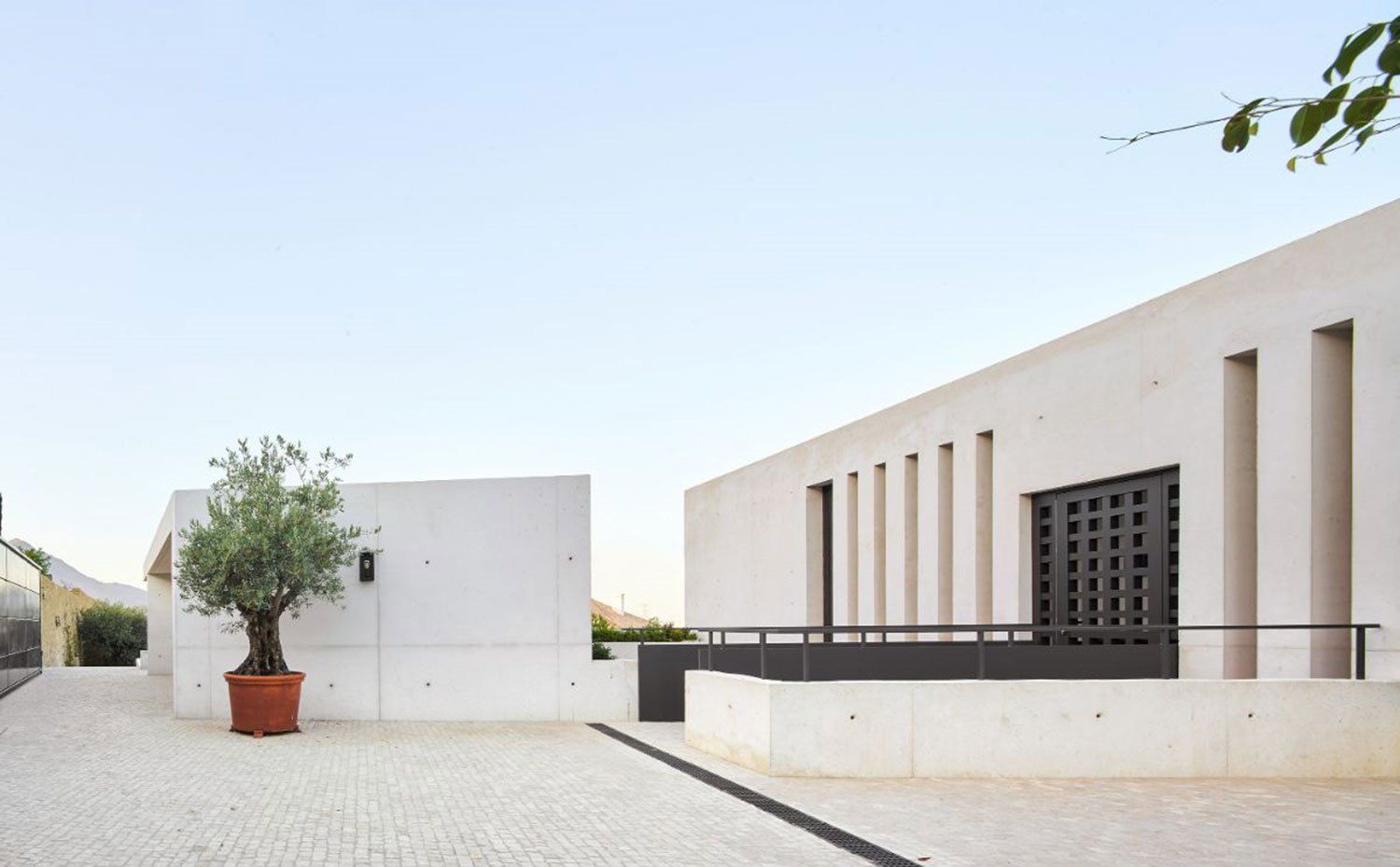 Zona de acceso principal a vivienda con pasarela y puerta de hierro, fachada con aperturas verticales de gran altura