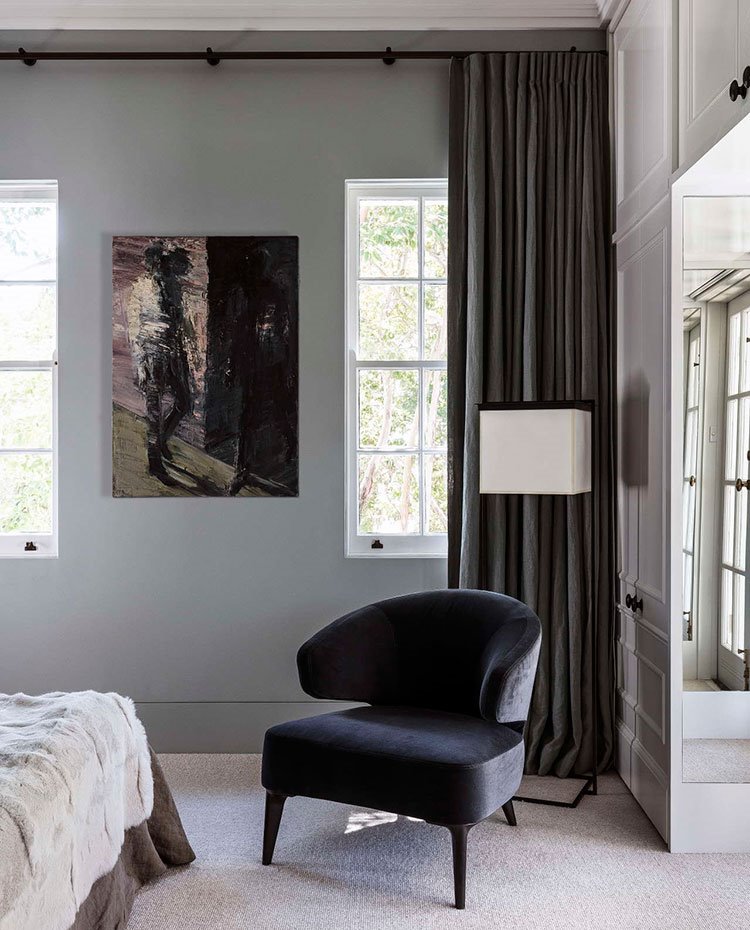 Sillón negro a los pies de la cama del dormitorio, con suelo enmoquetado, luminaria de pie junto a cortinas verdes y molduras en paredes