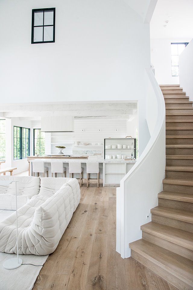 Escaleras desde salón a planta superior, pavimento de madera, sofás en blanco, cocina abierta con isla de trabajo