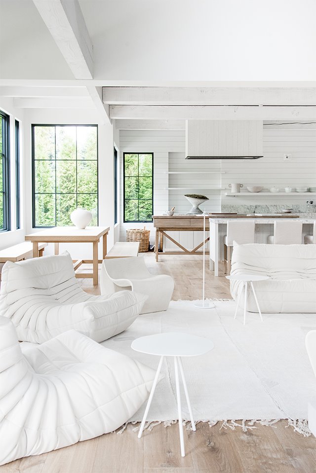 Salón con mobiliario en blanco, suelo de madera y comedor y cocina integrados, vistas al exterior de jardín