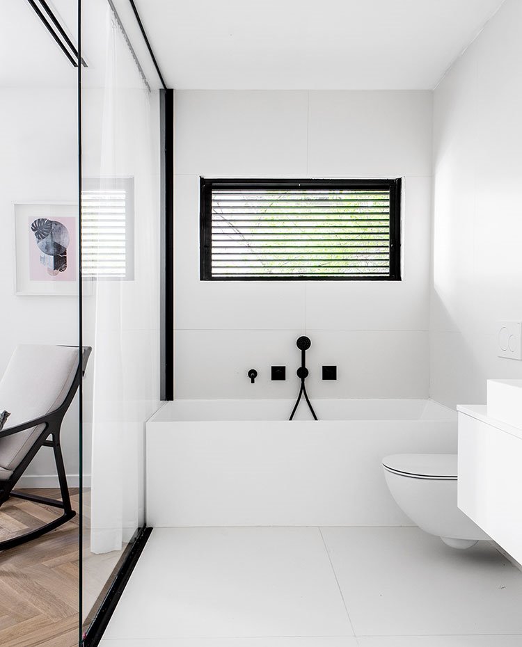 Cuarto de baño junto a habitación con cerramiento con tabique transparente, bañera, inodoro y lavamanos en blanco con grifería y perfilería en negro.