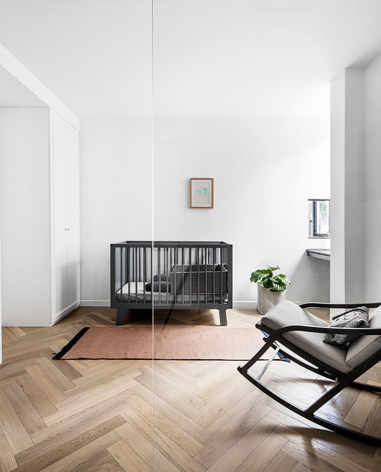 Cuna central en gris oscuro, alfombra marrón, mecedora con asiento en gris claro, armario blanco, suelo de madera, hija de cristal transparente