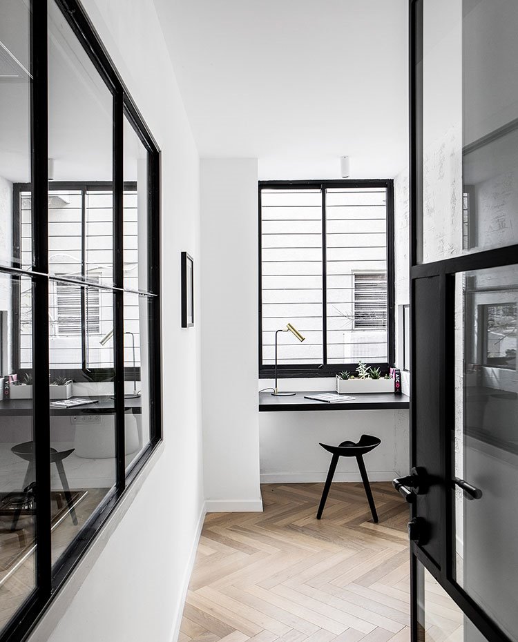 Rincón de trabajo con estructura de madera bajo ventana, taburete bajo negro, puerta de entrada y ventana a estancia continua de cristal con perfilería en negro