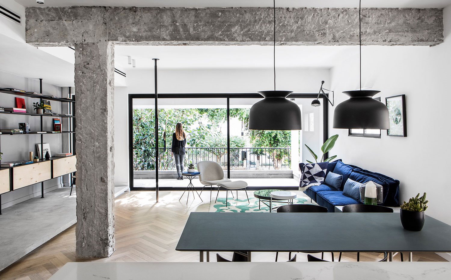 Vista de salón desde cocina con luminarias suspendidas en negro, vigas en cemento sin pulir y gran apertura a exterior