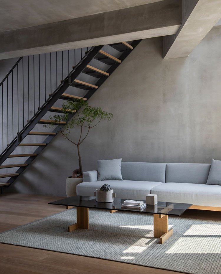 Salón bajo escalera con sofá gris, mesa con sobre de cristal y base de madera sobre alfombra gris, vigas de hormigón bajo techo