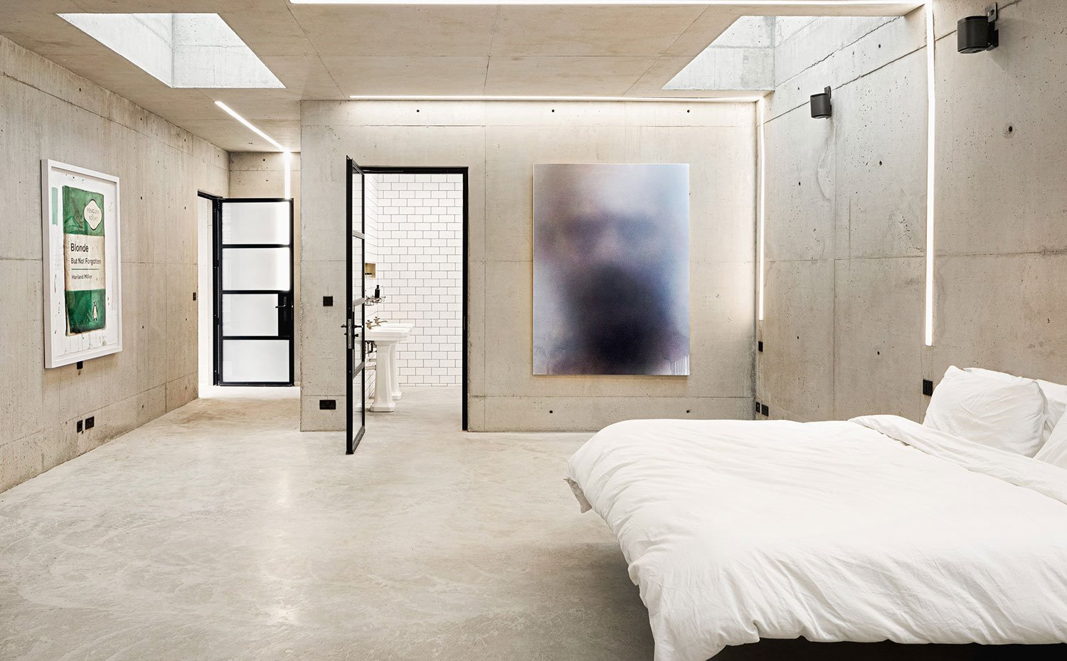 Dormitorio con revestimiento de hormgión, lencería de cama en blanco, puerta perfilería negra y cristal translúcido, claravboya en techo