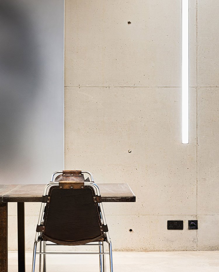 Mesa y silla de comedor con revestimiento de hormigón, lumiarias en estrechas aperturas en pared y cerramientos en cristal