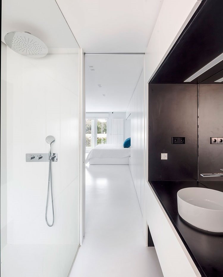 Baño-junto-a-dormitorio-en-línea-con-zona-de-almacenaje,-con-zona-de-tocador-en-negro,-con-lavamanos-en-blanco,-como-revestimiento-y-suelo,-y-grifería-de-ducha-encastrada-