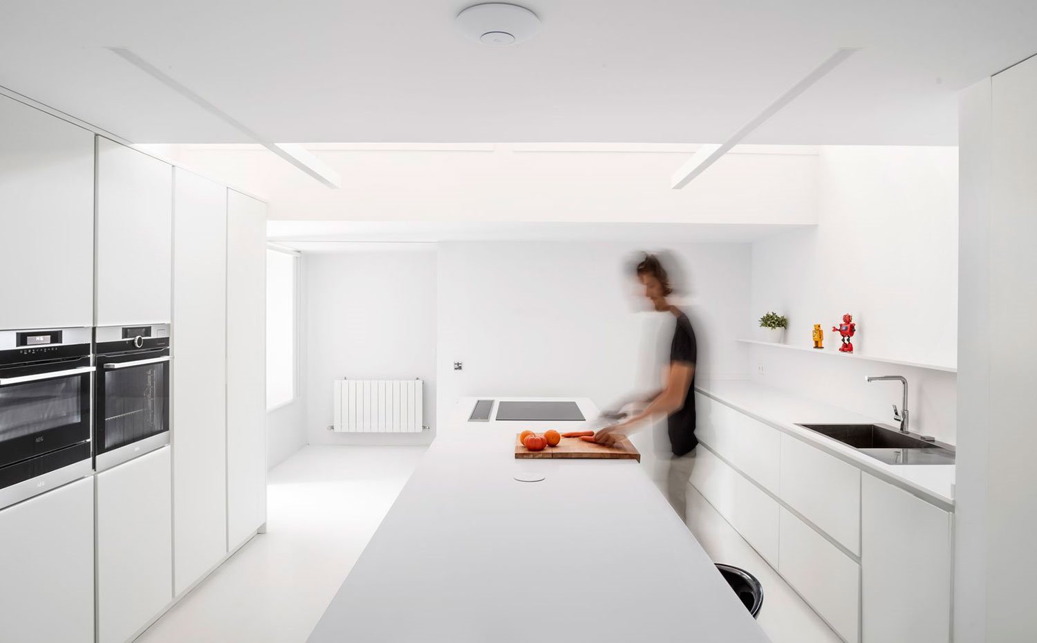 Cocina-con-isla-central-de-trabajo-y-zona-de-office-con-taburete-negro,-mobiliario-todo-en-blanco,-suelo,-paredes-y-techo-también-blanco,-detalles-de-grifería-y-electrodomésticos-en-acabado-cromado.-