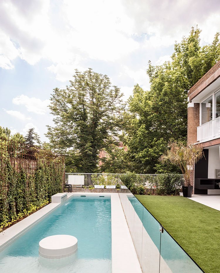 Piscina-exterior-alargada,-barra-protectora-en-cristal-transparente,-zona-ajardinada-con-cesped-frente-a-la-vivienda-de-ladrillo-visto-y-barandilla-blanca