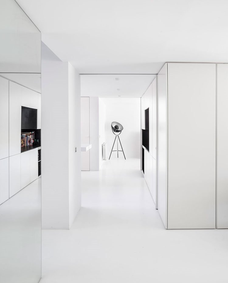 Zona de paso hacia cocina con suelo y mobiliario en blanco, pared revestida de espejo y luminaria de pie de carácter teatral