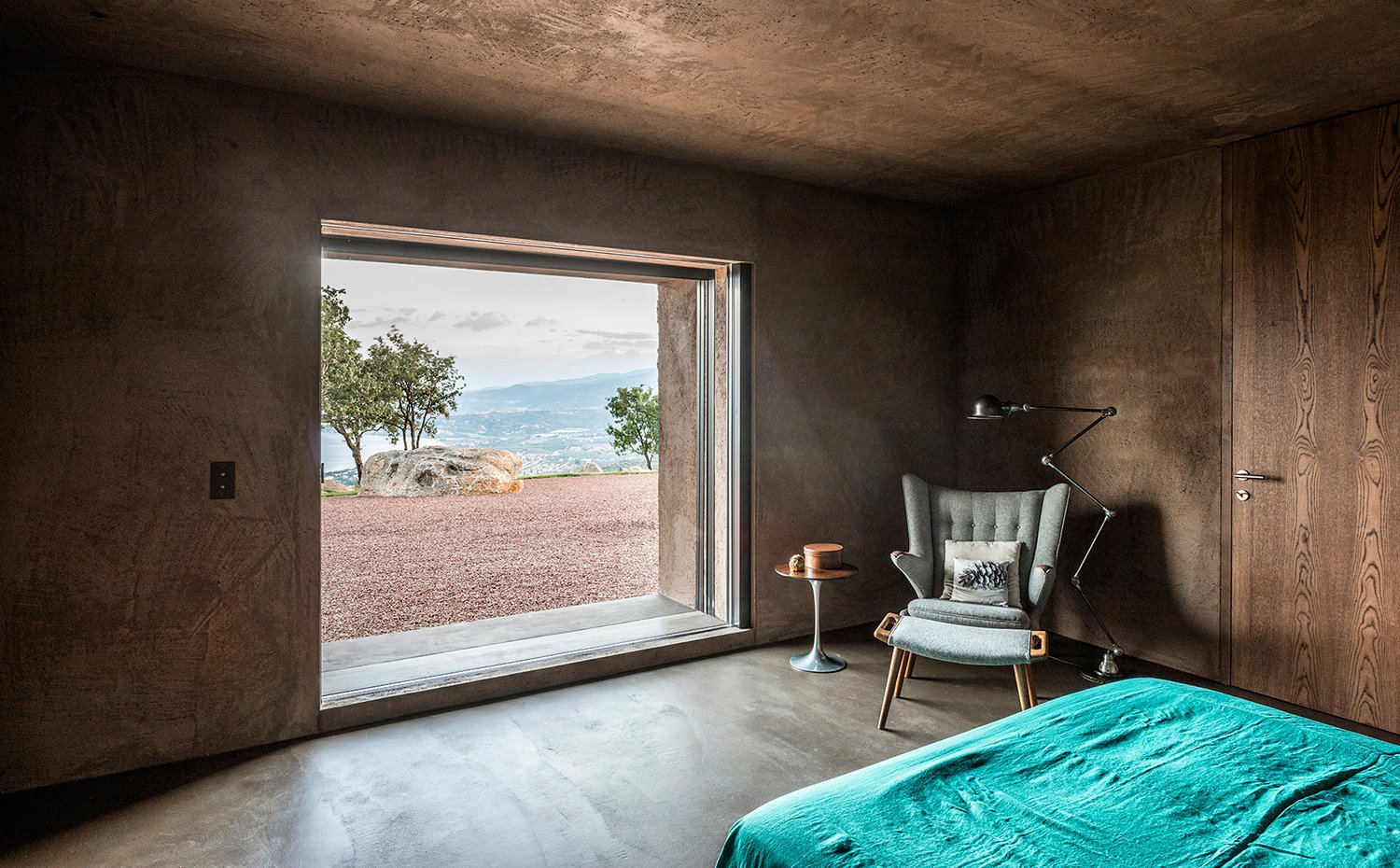 Dormitorio con estructura arcillosa, puerta de madera, gran ventana a exterior, zona de lectura y cama