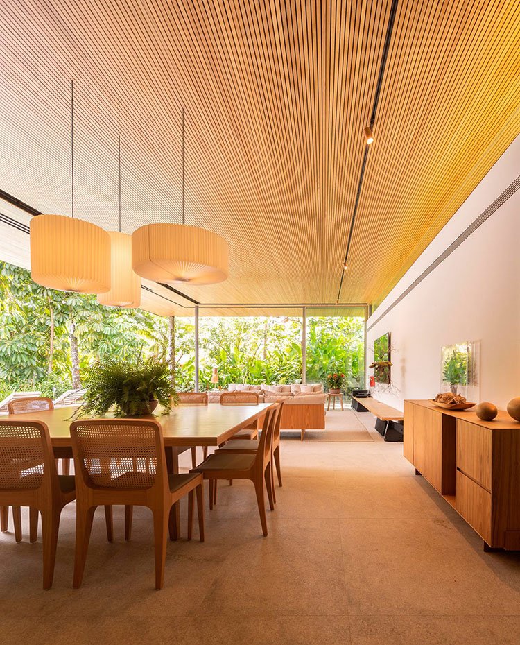 Comedor con gran mesa cuadrada, tres luminarias sobre ella y mueble contenedor, todo de madera