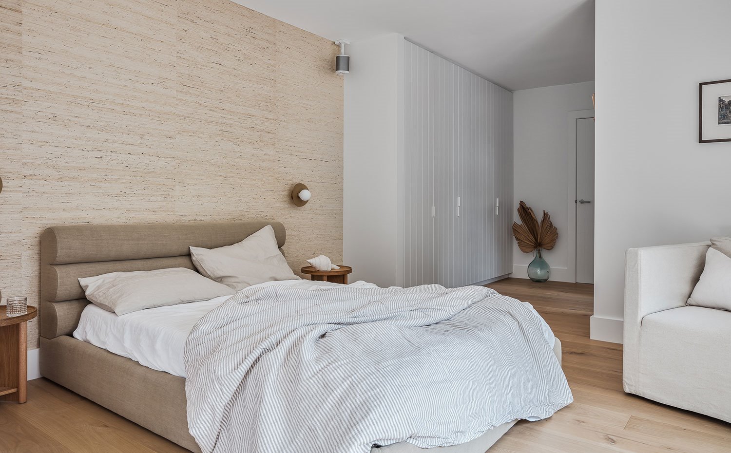 Dormitorio con cama tapizada en tejido natural color café, armarios blancos, sillón crudo, jarrón de cristal con hoja seca