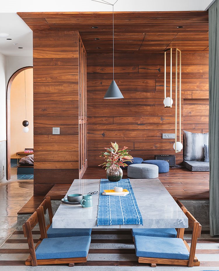 Zona de comedor con luminaria suspendida en gris, sillas en madera y cojín azul y al fondo, la zona de televisión